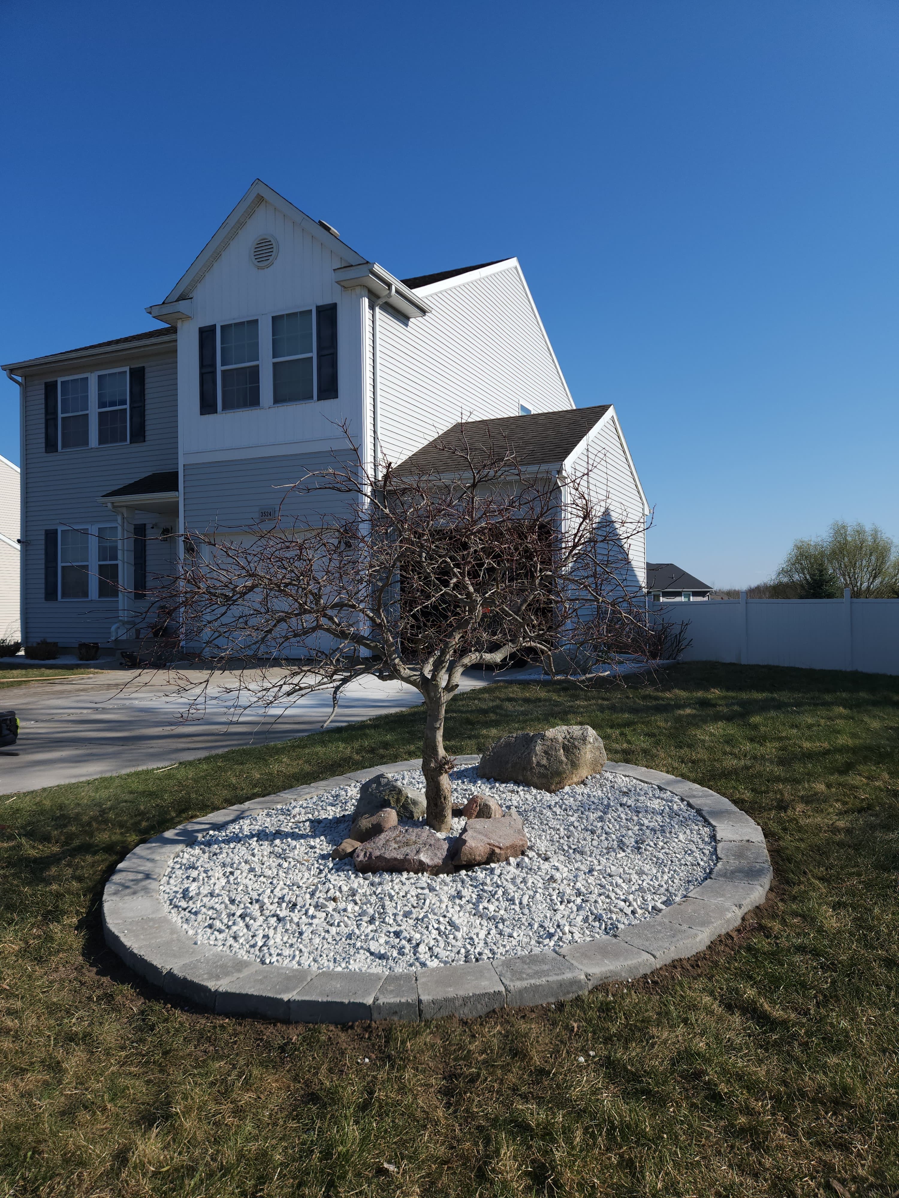 Circular paver-edged front-yard bed with white rock, an ornamental tree, and a boulder accent