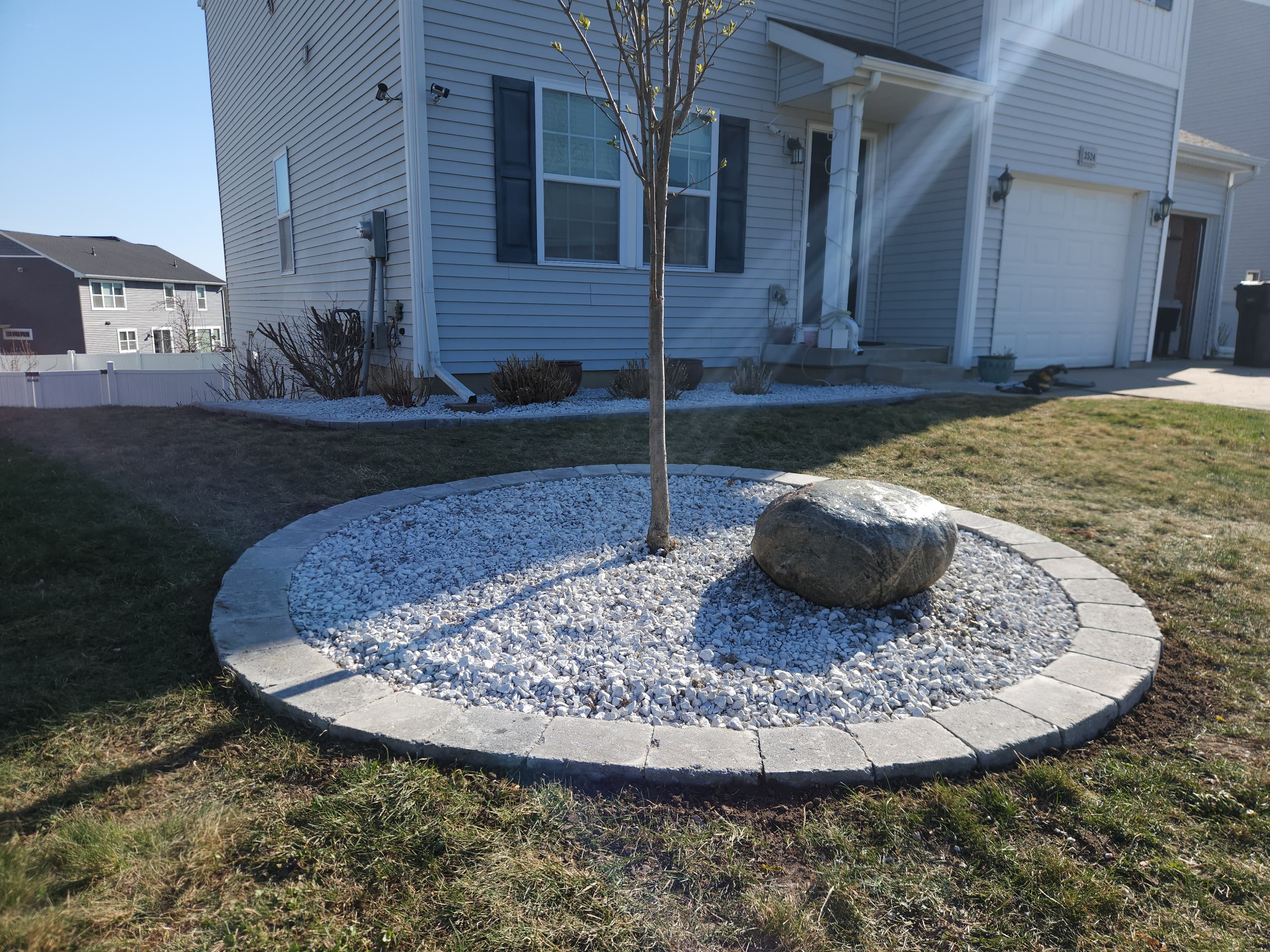 Circular paver-edged front-yard island bed with white rock and a slender ornamental tree
