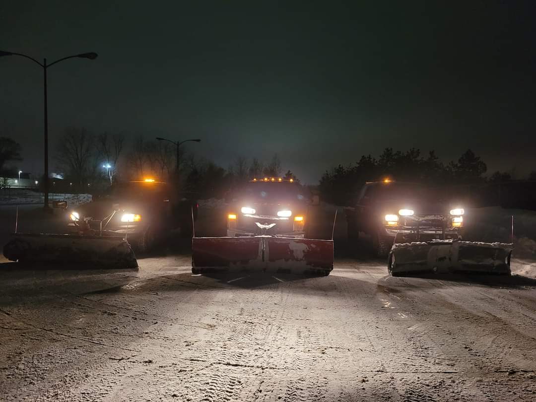 Three Bernal plow trucks lined up at night on snowy pavement with plows lowered