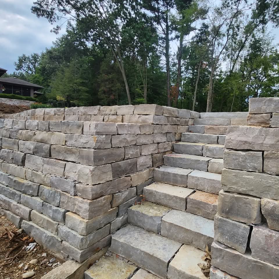 Tall rustic natural stone retaining wall with a wide stone staircase in a wooded backyard