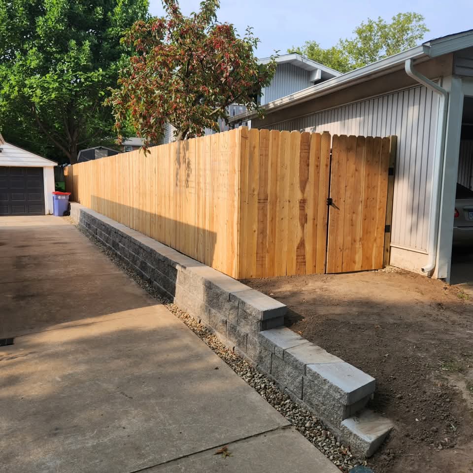 Low gray block retaining wall with cap stones along a driveway with a new cedar privacy fence and gate on top