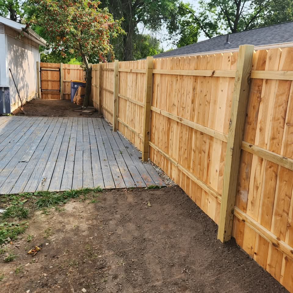 New cedar privacy fence sections along a property line with a weathered wood deck behind and bare dirt in the foreground