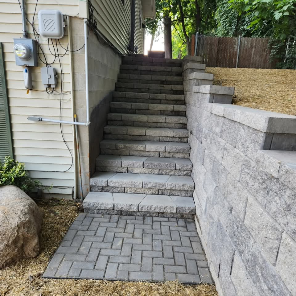Gray block retaining wall with integrated steps and herringbone paver landing at the base next to a house