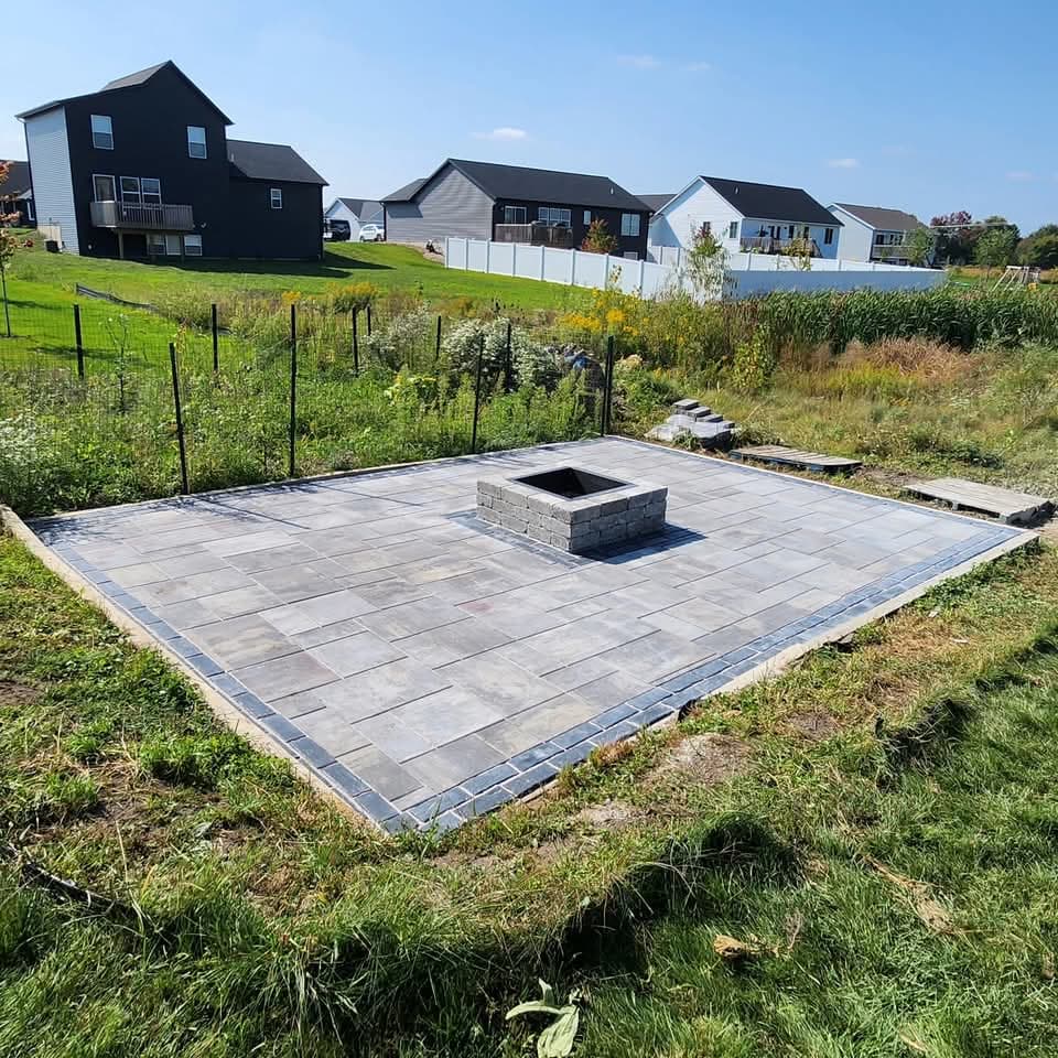 Gray rectangular paver patio with a central square stone fire pit in an open backyard