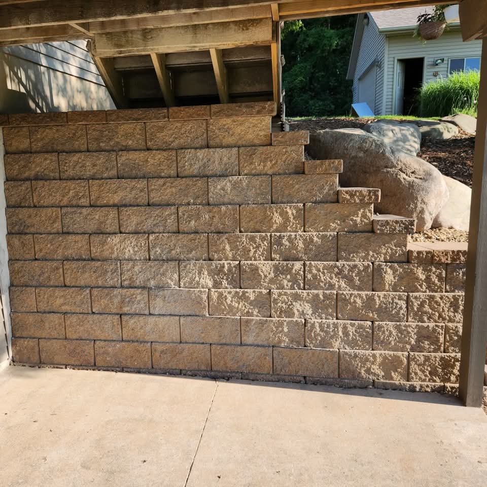 Tall stepped block retaining wall under a raised deck with a boulder accent viewed from the concrete pad below