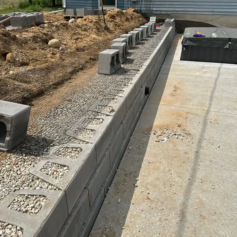 Concrete block retaining wall under construction with rebar grid and gravel base next to an existing concrete pad and hot tub