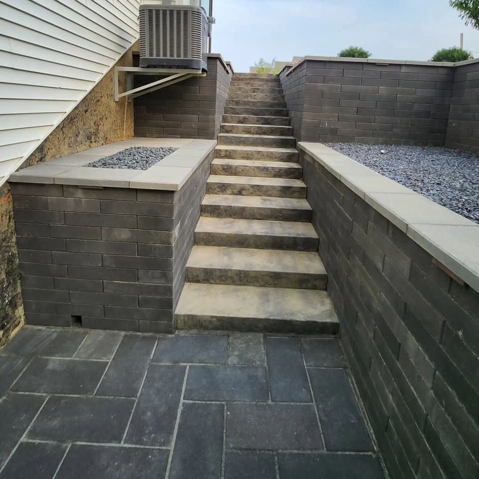 Dark block retaining walls on both sides of a sunken area with integrated block steps and light cap stones, gravel bed alongside a white house with an AC unit