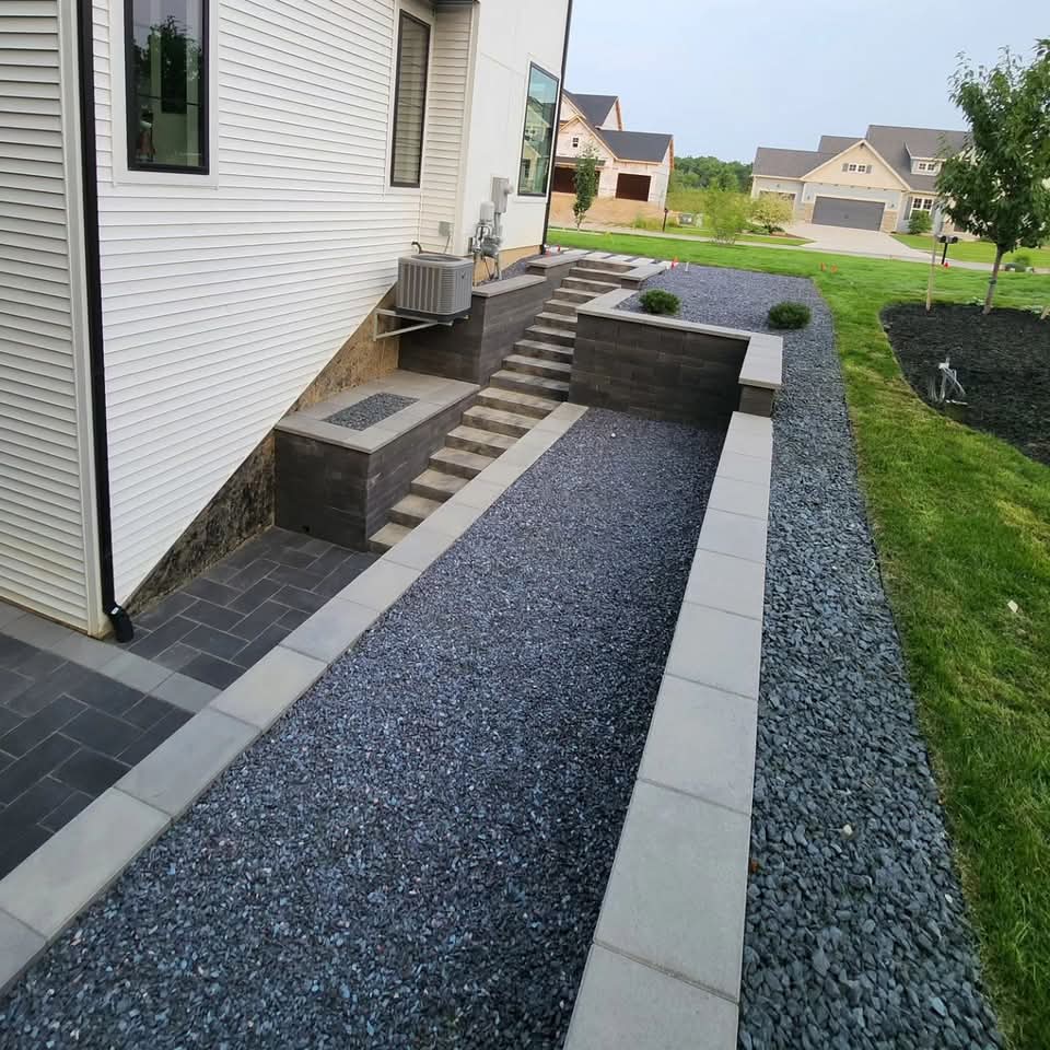 Expansive dark herringbone paver patio behind a modern two-story white house with a second-story balcony, wood-post covered area, and block retaining wall with gravel bed