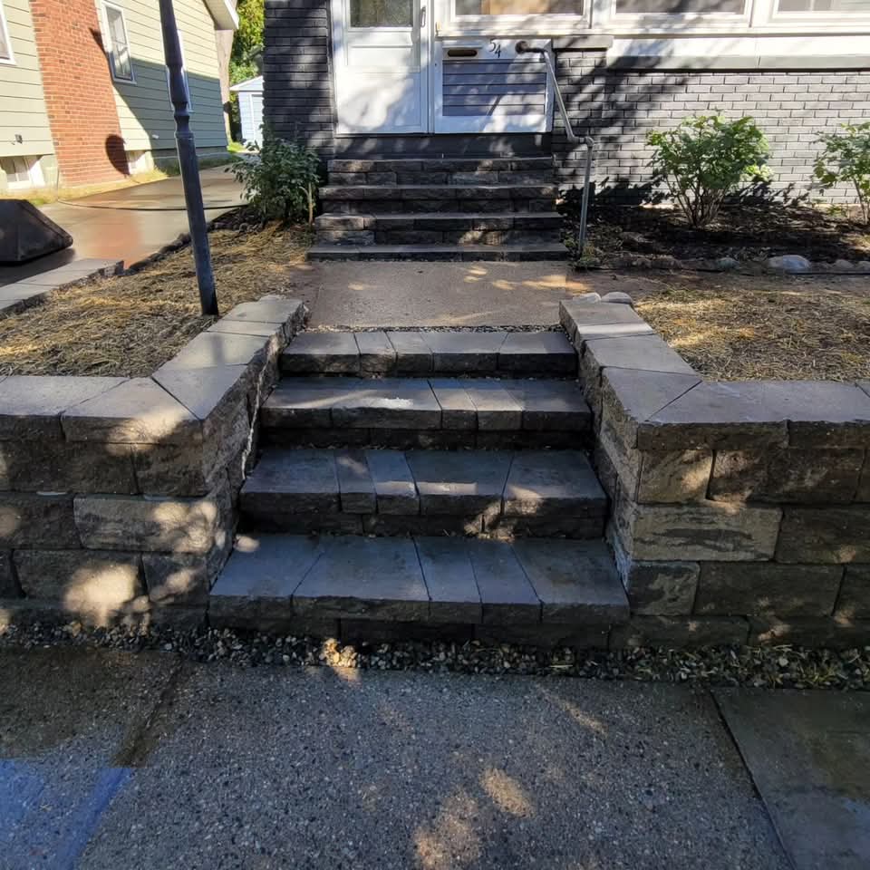 Front of a gray painted brick house with new block retaining wall and integrated block steps from the sidewalk to the front door, metal handrail, lamp post, and mulch beds with shrubs
