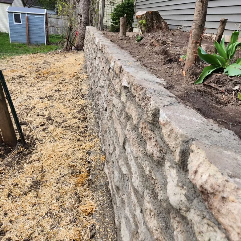 Two-tier block retaining wall with integrated steps from the sidewalk up to the front door of a gray painted brick house, metal handrail, and straw-covered seed on both sides