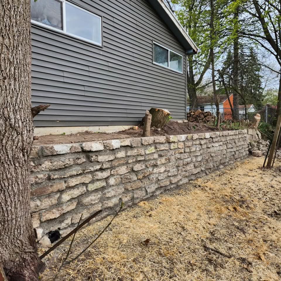 Long natural stone retaining wall in a residential backyard with straw-covered new seed on one side and bare dirt with plantings on the other, blue shed in the background