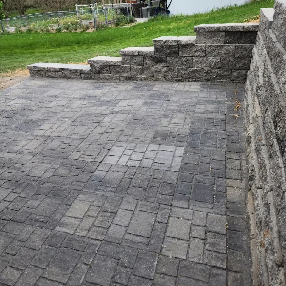 Rough-cut natural stone retaining wall along a residential side yard next to a gray house with siding, straw-covered seed, cut tree stumps, and a wood pile behind