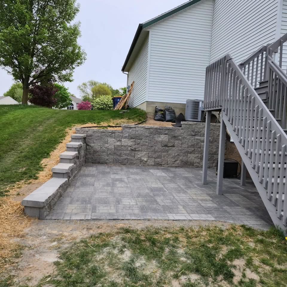 Sunken paver patio with textured block retaining wall and integrated block steps, green hillside lawn above, and chain-link fence in the background