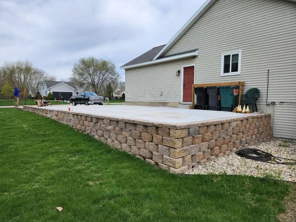 Two-tier curved gray-green block retaining wall with dark mulch fill and cap stones along the foundation of a green-sided house with a stone column porch, dark mulch groundcover below, and mature trees overhead