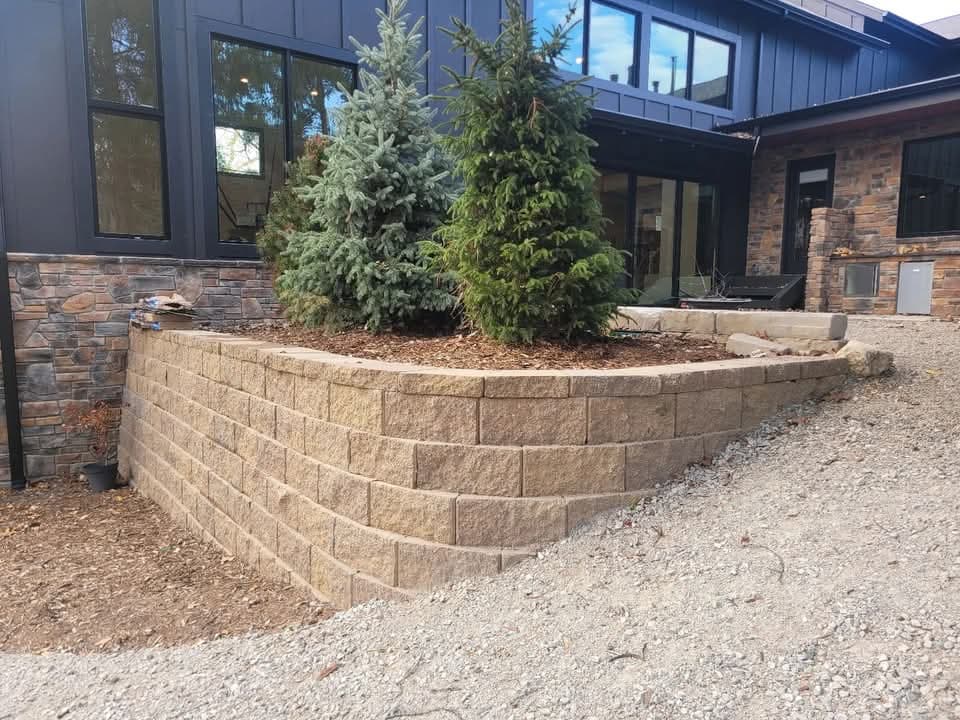 Brown block retaining wall supporting a raised concrete slab patio behind a gray-sided house with a red door, trash bins in a wood-framed shelter, green lawn below, and crew members with equipment at the far end