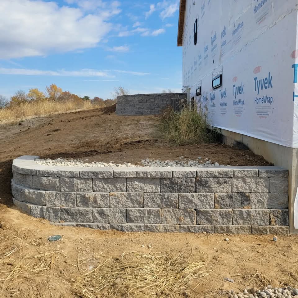 Curved gray block retaining wall at the foundation of a new construction home wrapped in Tyvek house wrap, white river rock along the wall top, bare dirt hillside, and autumn trees in the background