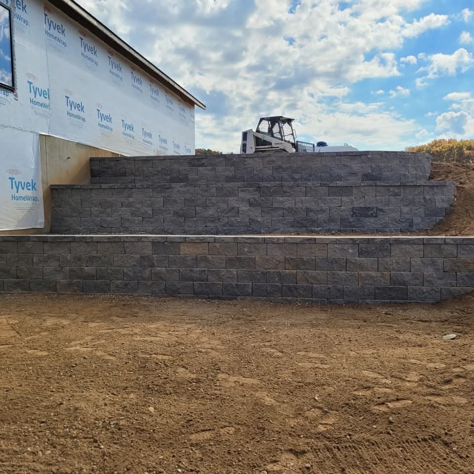 Two-tier dark charcoal block retaining wall at a new construction site with Tyvek house wrap visible, a skid-steer loader on the upper level, bare dirt grading, and an autumn sky