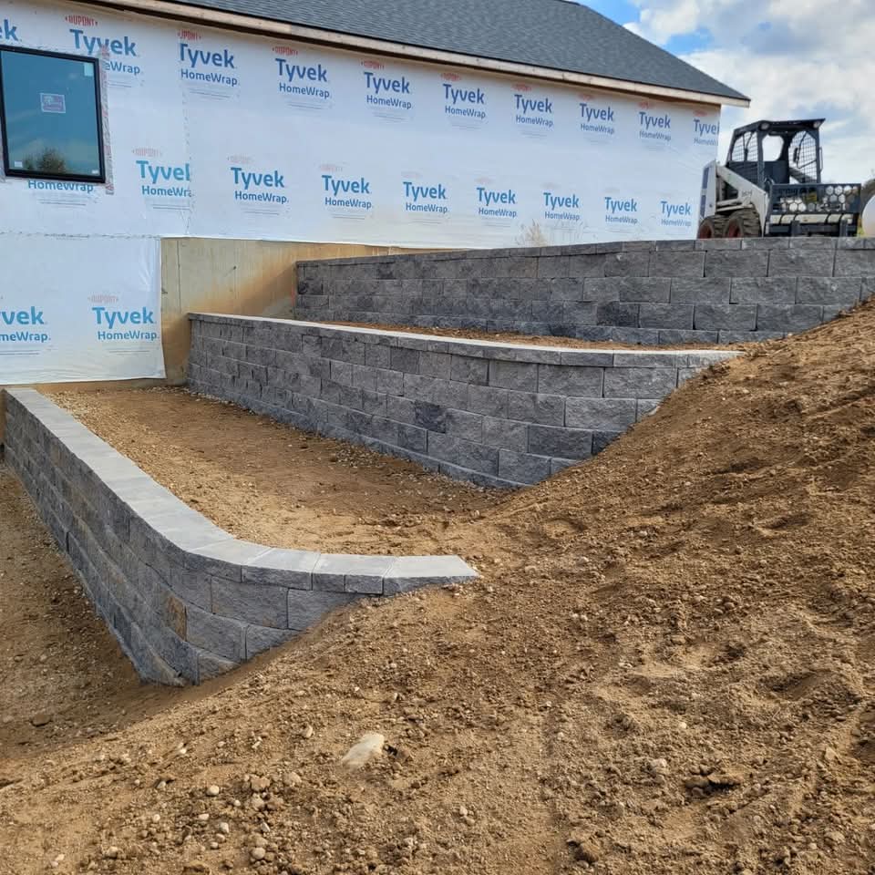 Two-tier dark charcoal block retaining wall with light cap stones at a new construction site, Tyvek house wrap on the wall behind, a skid-steer on the hillside above, and bare dirt grading