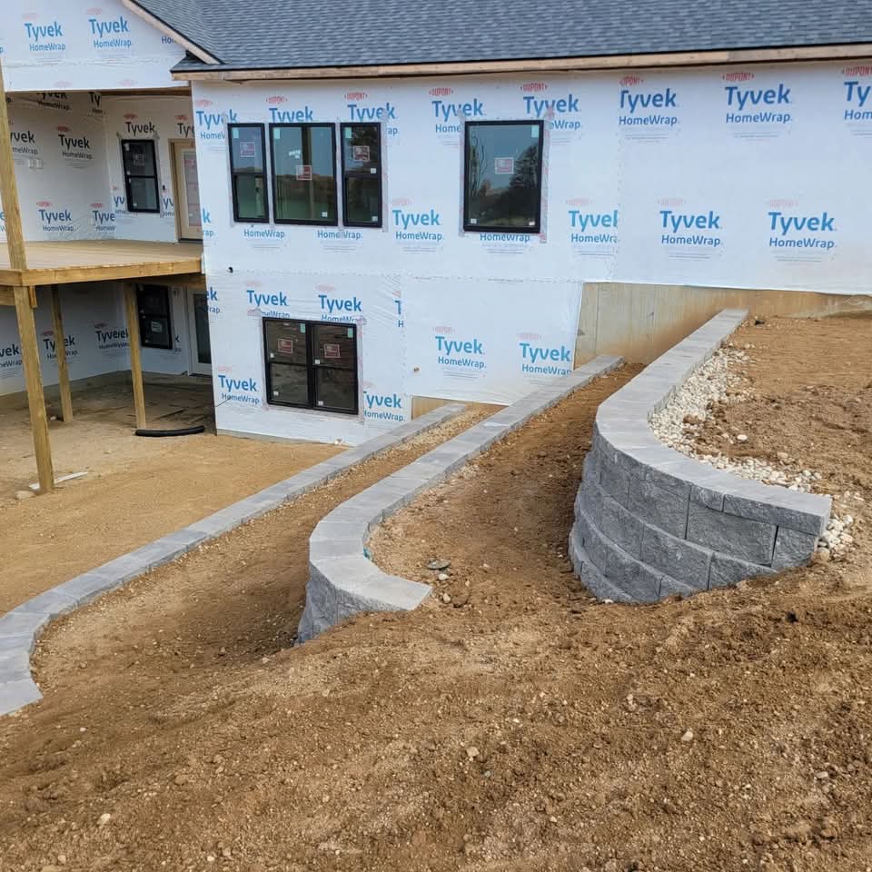 Curved gray block retaining wall with light cap stones wrapping around the foundation of a new construction home with Tyvek house wrap, white gravel drainage bed, and bare dirt lot