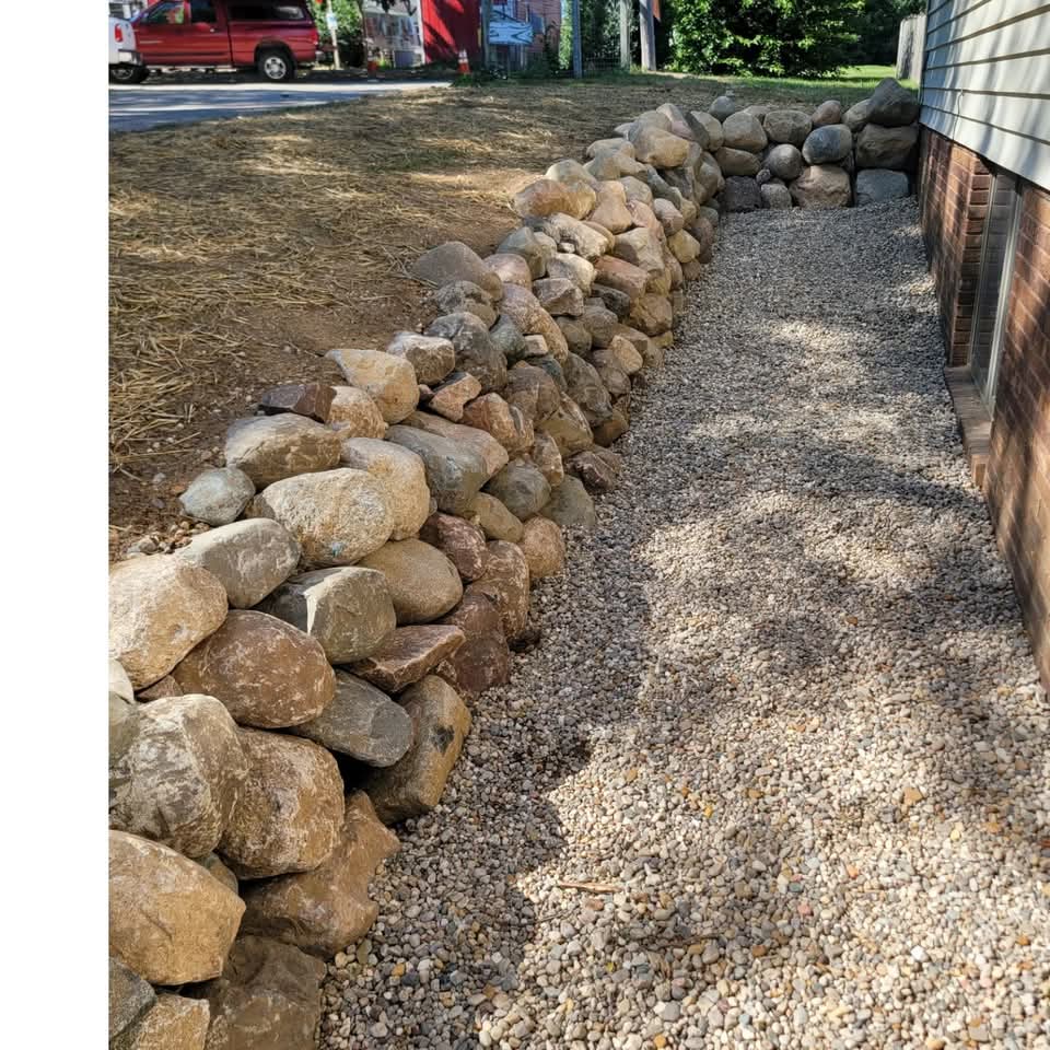 Natural fieldstone and boulder retaining wall along the side of a house with white siding and a brick foundation, gravel drainage bed between the wall and house, and straw-covered seed on the yard