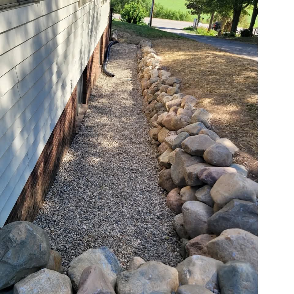 Natural fieldstone and boulder retaining wall along the side of a house with white siding and brick foundation, gravel bed with a downspout, straw-covered seed, and a rural road in the background