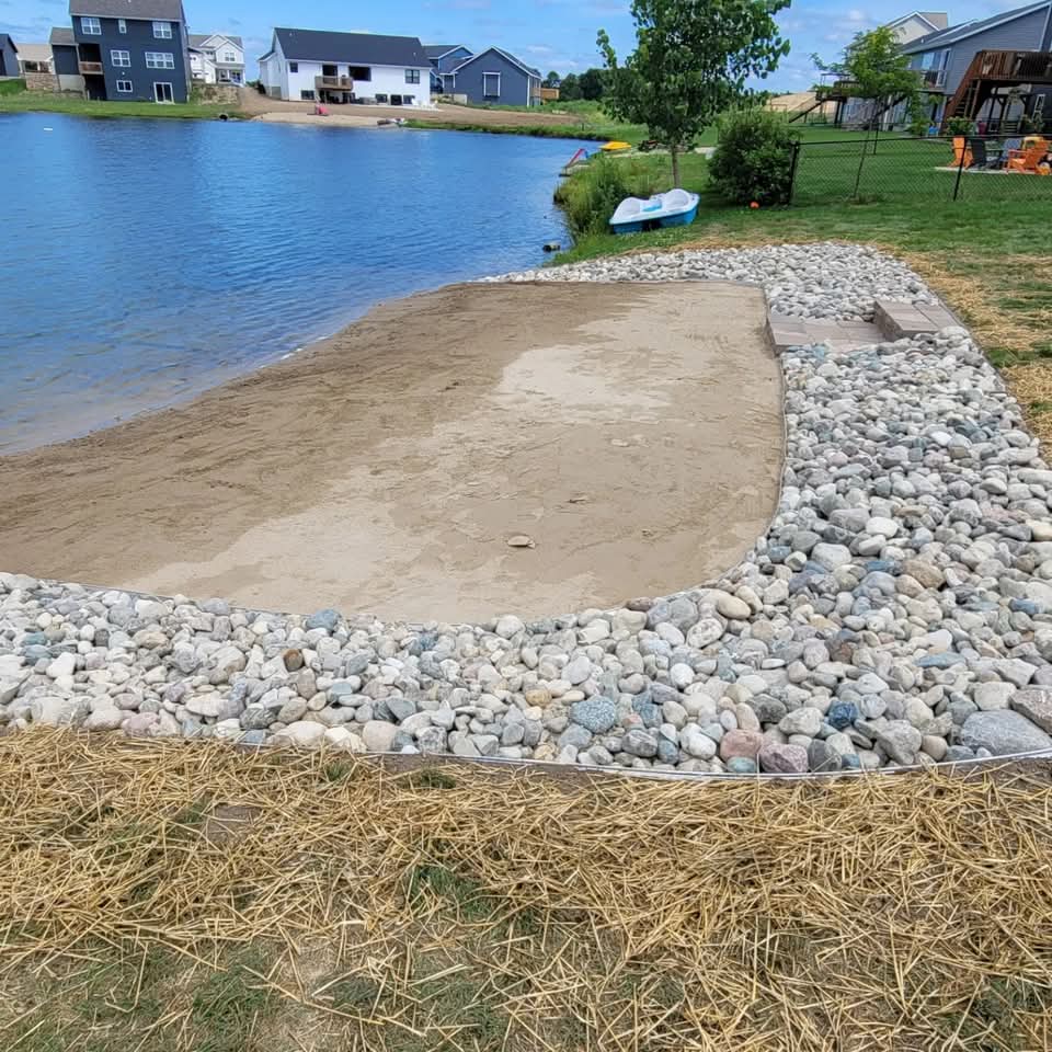 Lakefront residential property with a sand beach area bordered by large river rock, metal landscape edging, straw-covered new seed, and a pond with houses and a pedal boat visible across the water