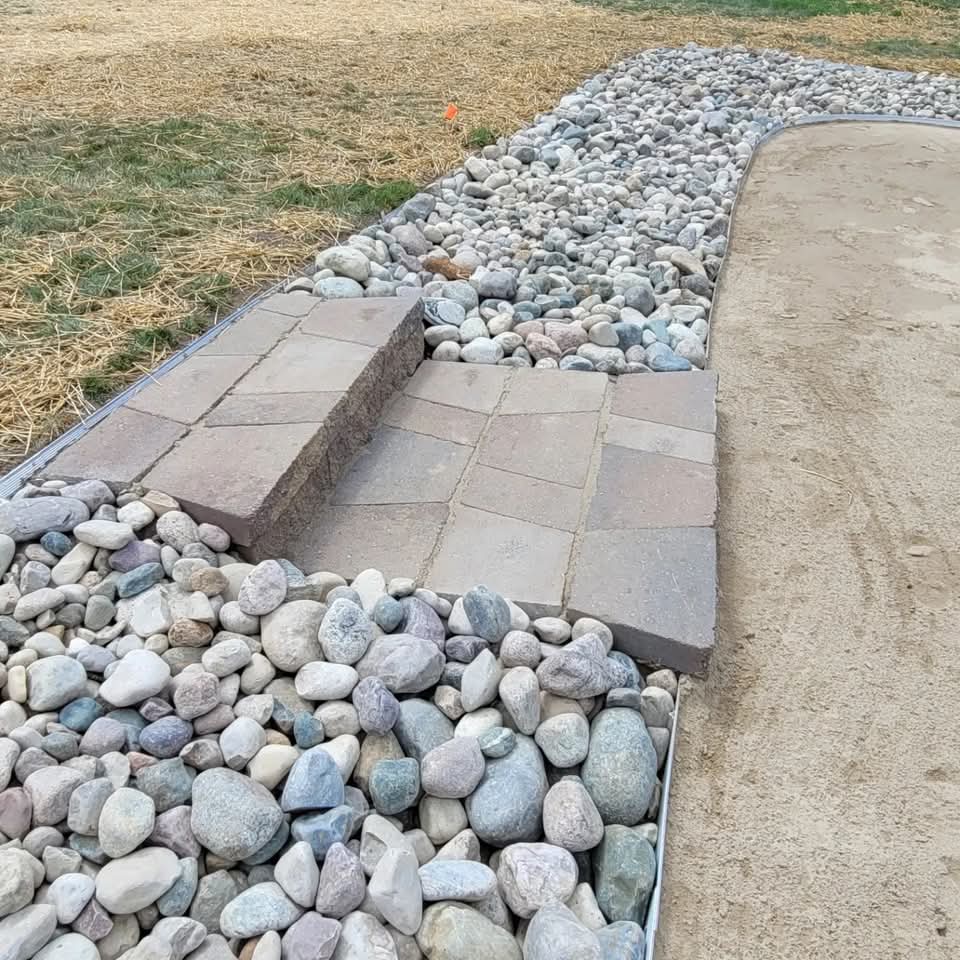 Close-up of brown paver steps with river rock on both sides and metal landscape edging, leading from a straw-covered lawn down to a sand beach area on a lakefront property