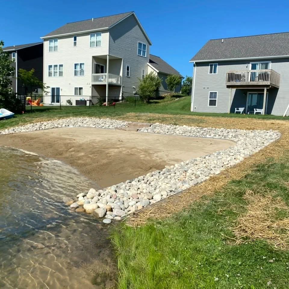 Lakefront sand beach area bordered by large river rock viewed from the water's edge, with two-story houses with second-floor balconies behind and straw-covered new seed on the lawn