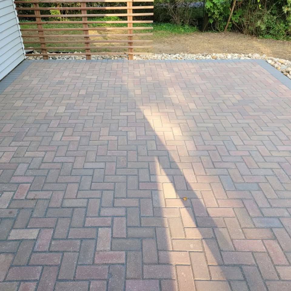 Red-brown herringbone paver patio in a backyard with a horizontal wood slat privacy fence, river rock border along the edges, and shrubs behind the fence