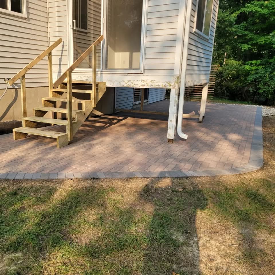 Red-brown herringbone paver patio beneath a raised deck with new wood stairs and railings, white downspout, gray soldier-course paver border, and a grass yard in the foreground