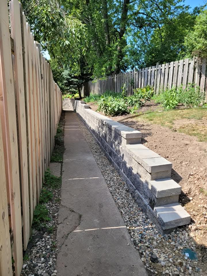 Dark charcoal textured block retaining wall with a drain pipe opening built between concrete foundation walls and outbuildings, large evergreen trees behind, bare dirt and debris in the foreground
