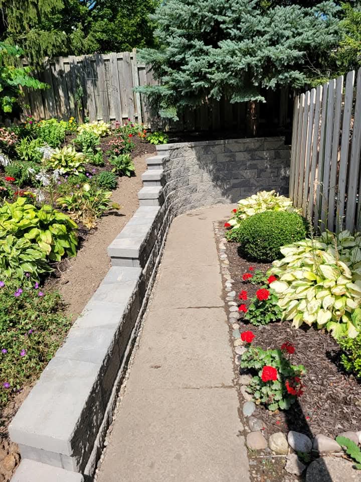 Gray block retaining wall with cap stones along a narrow walkway between a cedar fence and a raised planting bed, view through a fence opening showing flowering plants and an evergreen tree, gravel drainage strip along the base