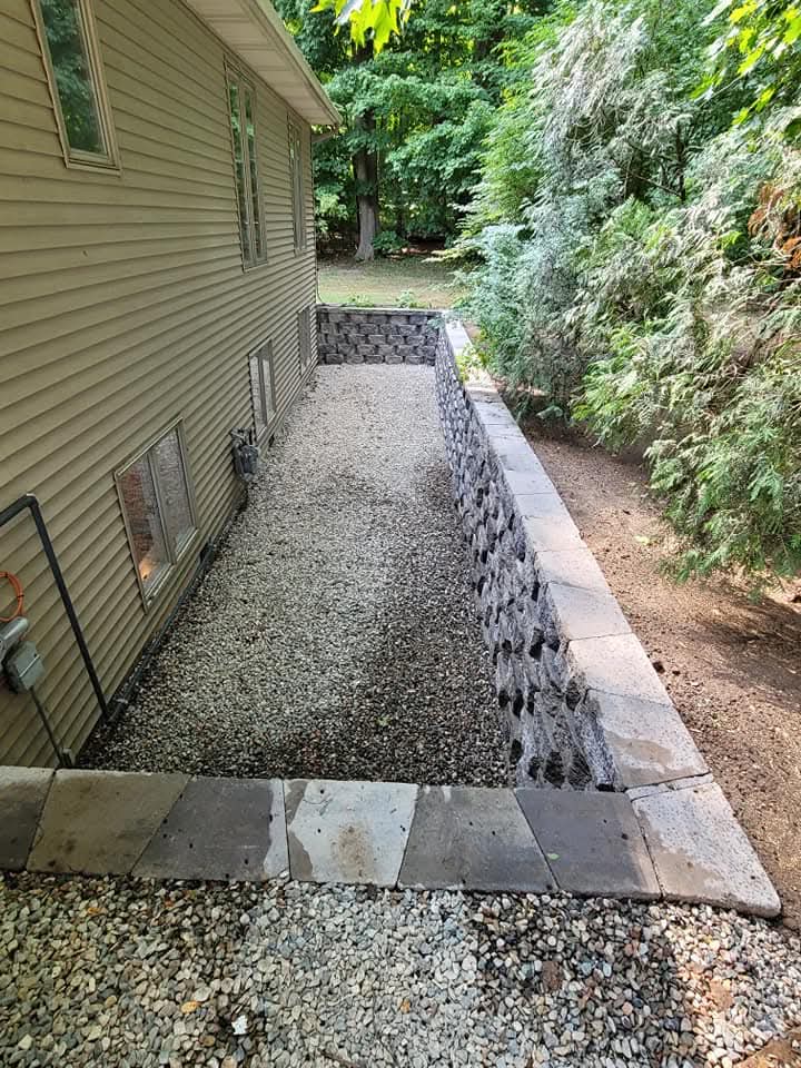 Concrete walkway with a gray block retaining wall and cap steps on one side and planting beds on both sides featuring hostas, red flowers, and green shrubs, block steps leading up to a cedar fence in the background