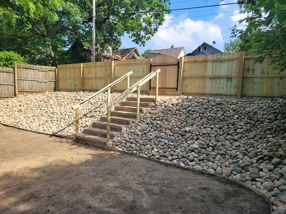 Backyard with a new cedar privacy fence, a river rock slope with wood stairs and a wood handrail leading up to a gate in the fence, bare dirt grading in the foreground, and large trees overhead