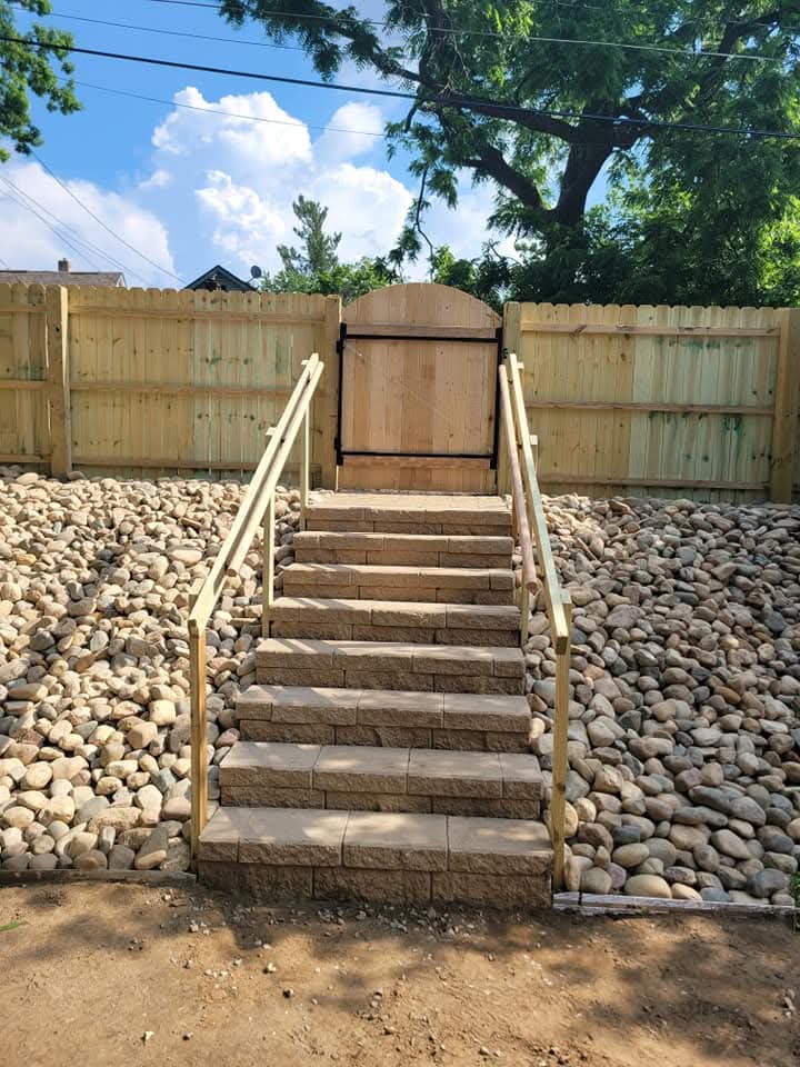 Block steps with paver treads and wood post-and-rail handrails on both sides leading up through a river rock slope to a cedar privacy fence with an arched gate at the top, mature trees overhead