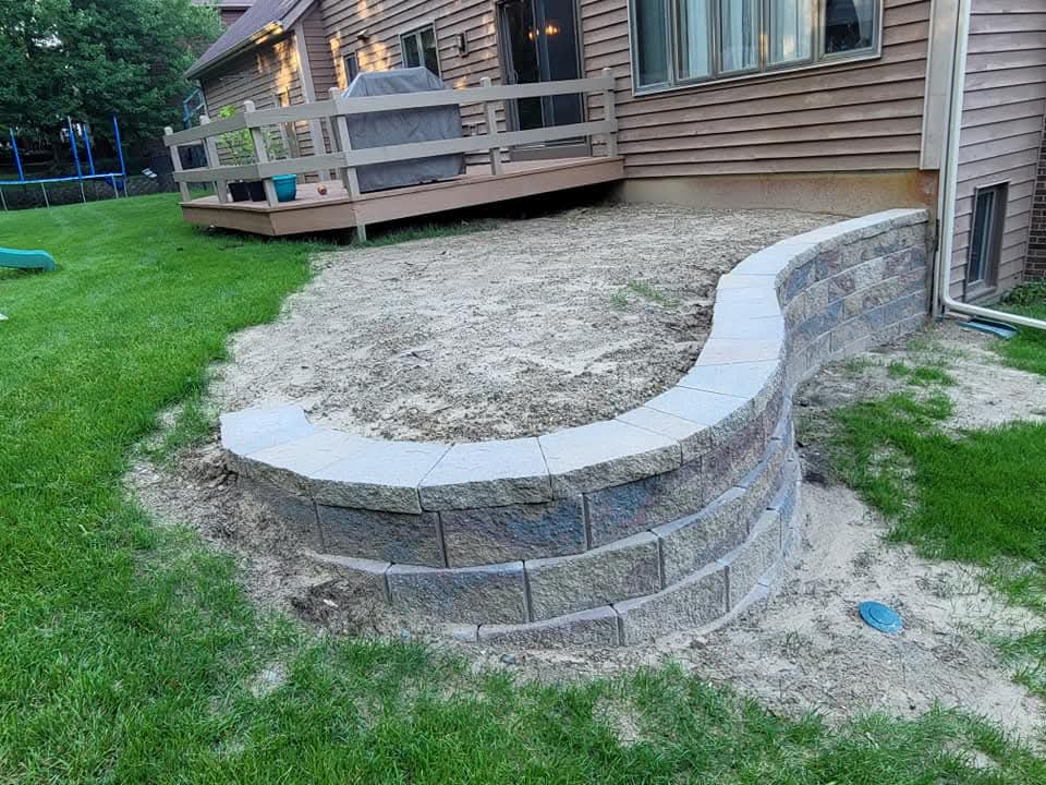 Curved gray block retaining wall with cap stones under construction behind a brown-sided house with a wood deck, bare sand fill inside the wall, green lawn and a trampoline in the background