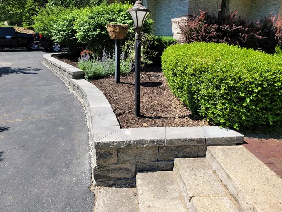Short gray block retaining wall with cap stones and a black lamppost along a curved asphalt driveway, dark mulch bed with rounded green shrubs and purple flowers, natural stone steps at the corner, brick house partially visible