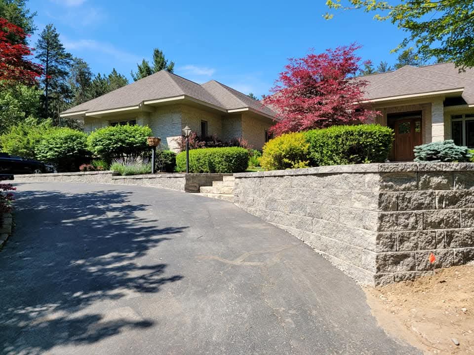 Long gray block retaining wall with cap stones along a curved asphalt driveway leading to a tan brick ranch-style home with manicured green shrubs, a red-leafed tree, and tall pine trees in the background