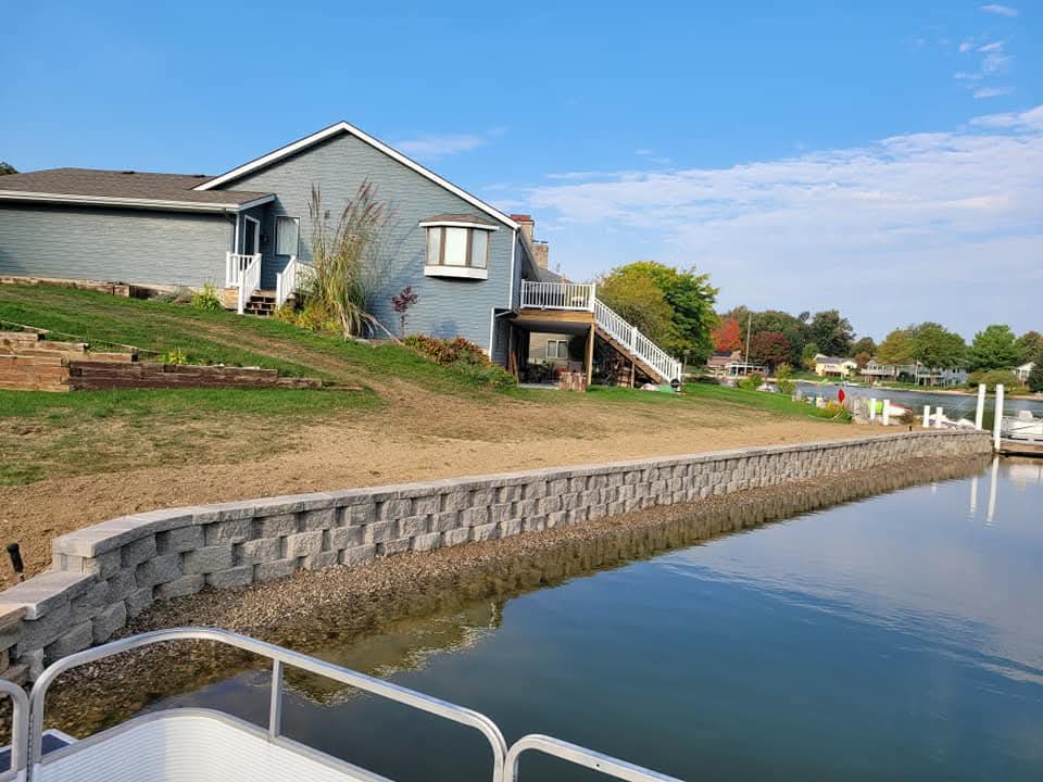 Gray block retaining wall with cap stones curving along a lakefront shoreline, a blue-sided house with white deck stairs on the green lawn above, tall ornamental grasses, straw-covered new seed on the slope, boats and docks on the water