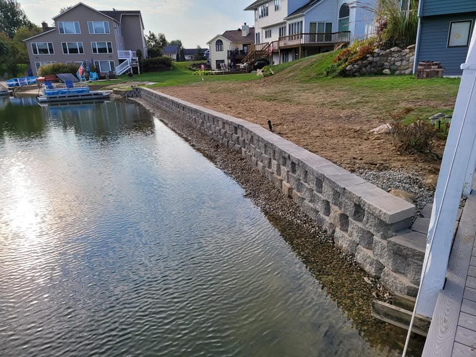 Long gray block retaining wall with cap stones along a lakefront channel, multiple two-story houses with green lawns on the slope above, a pontoon boat docked on the left, a natural boulder wall visible on a neighboring property