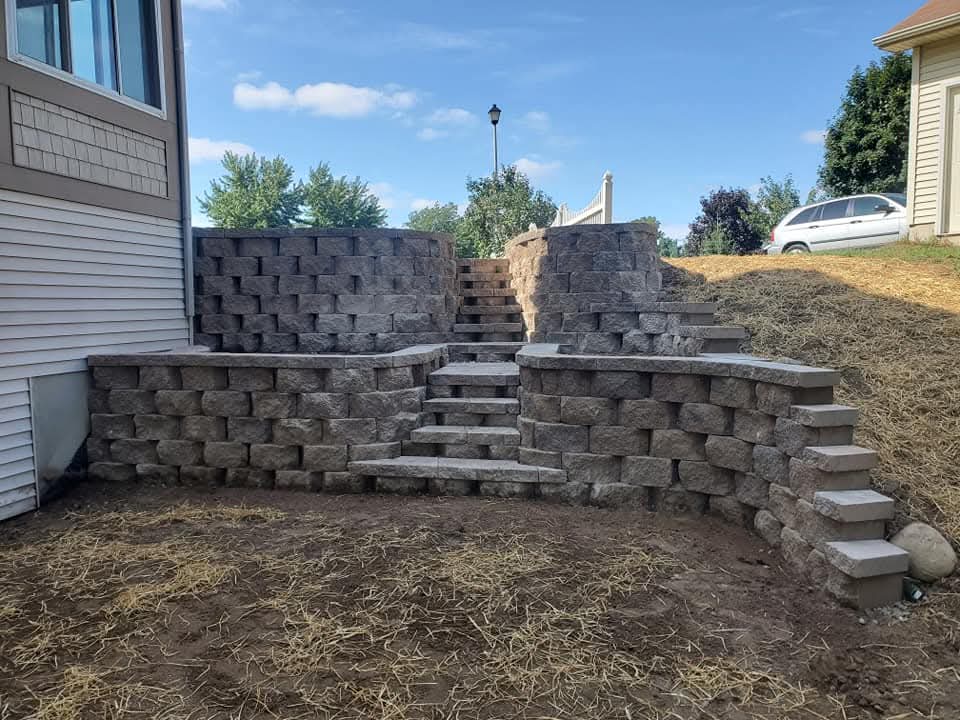 Multi-tier dark charcoal block retaining wall with two sets of block steps on a steep slope between two houses, straw-covered new seed on the hillside, a lamppost and white deck railing at the top, blue sky overhead