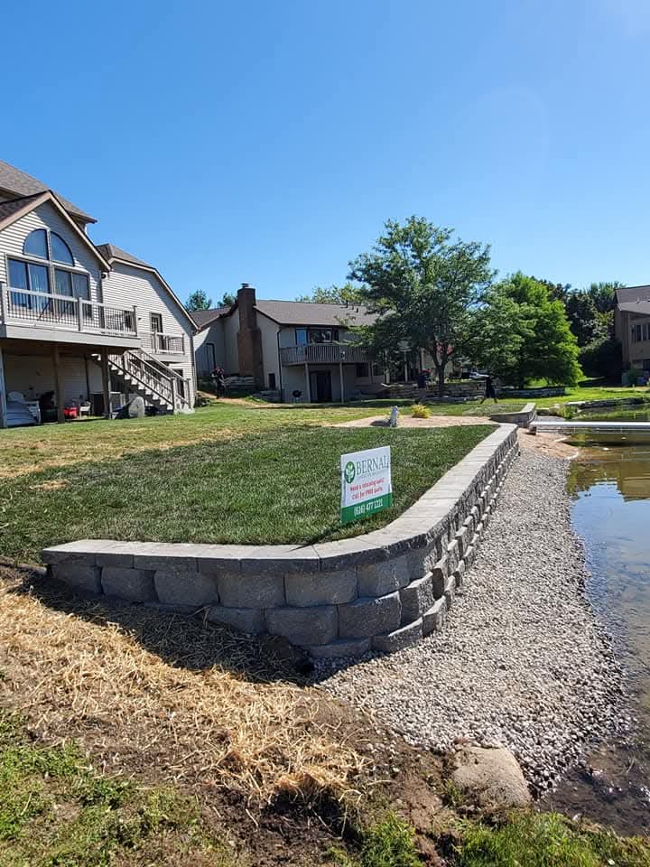 Gray block retaining wall with cap stones curving along a lakefront pond, a Bernal Landscape Management yard sign on the green lawn, a tan two-story house with a wood staircase and balcony above, neighboring homes across the water