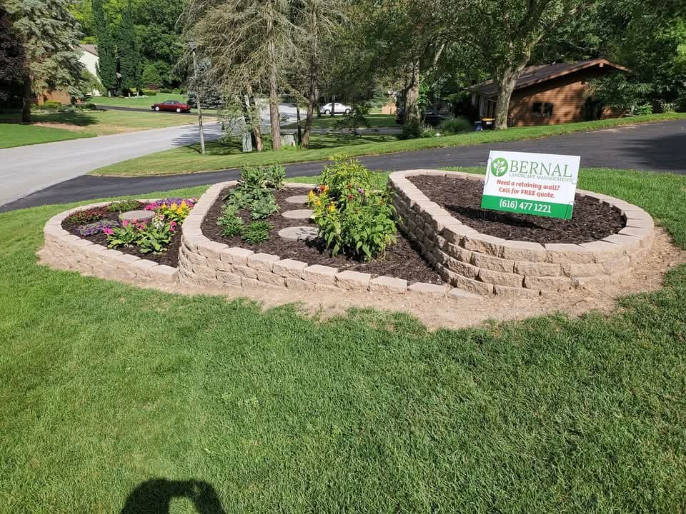 Decorative kidney-shaped block planter with two tiers of tan block walls, dark mulch and colorful flowering plants inside, a Bernal Landscape Management yard sign, green lawn surrounding, a residential street and brown building in the background