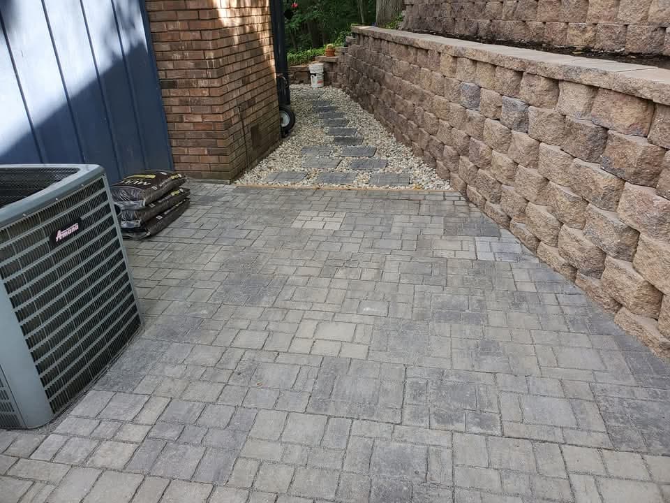 Gray paver patio surface along a tan block retaining wall next to a brick house, an air conditioning unit and bags of material on the left, river rock drainage strip between the wall and a narrow walkway, potted flowers on the wall cap