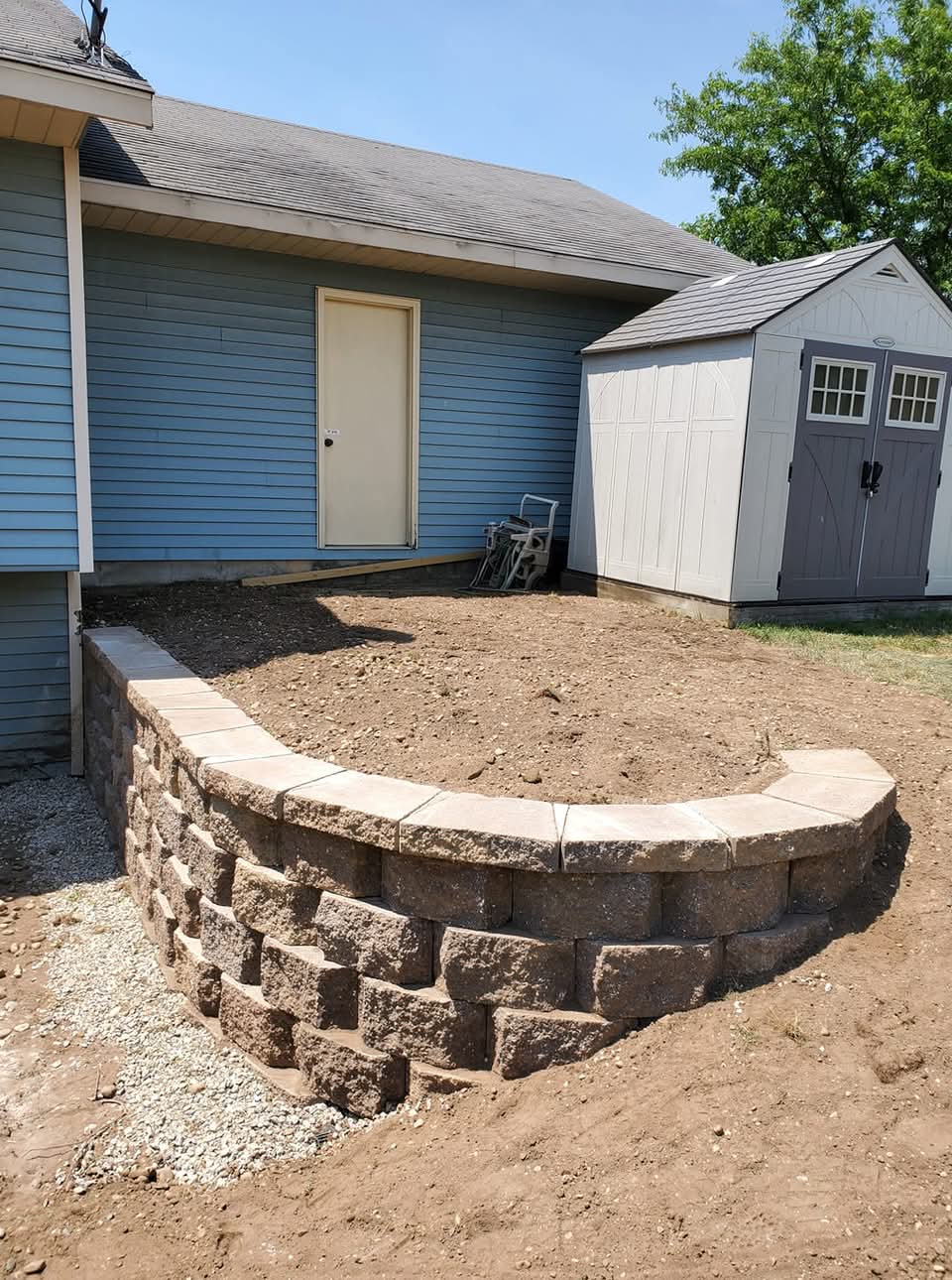 Curved block retaining wall with gravel fill creating a raised bed area next to a blue house and a storage shed