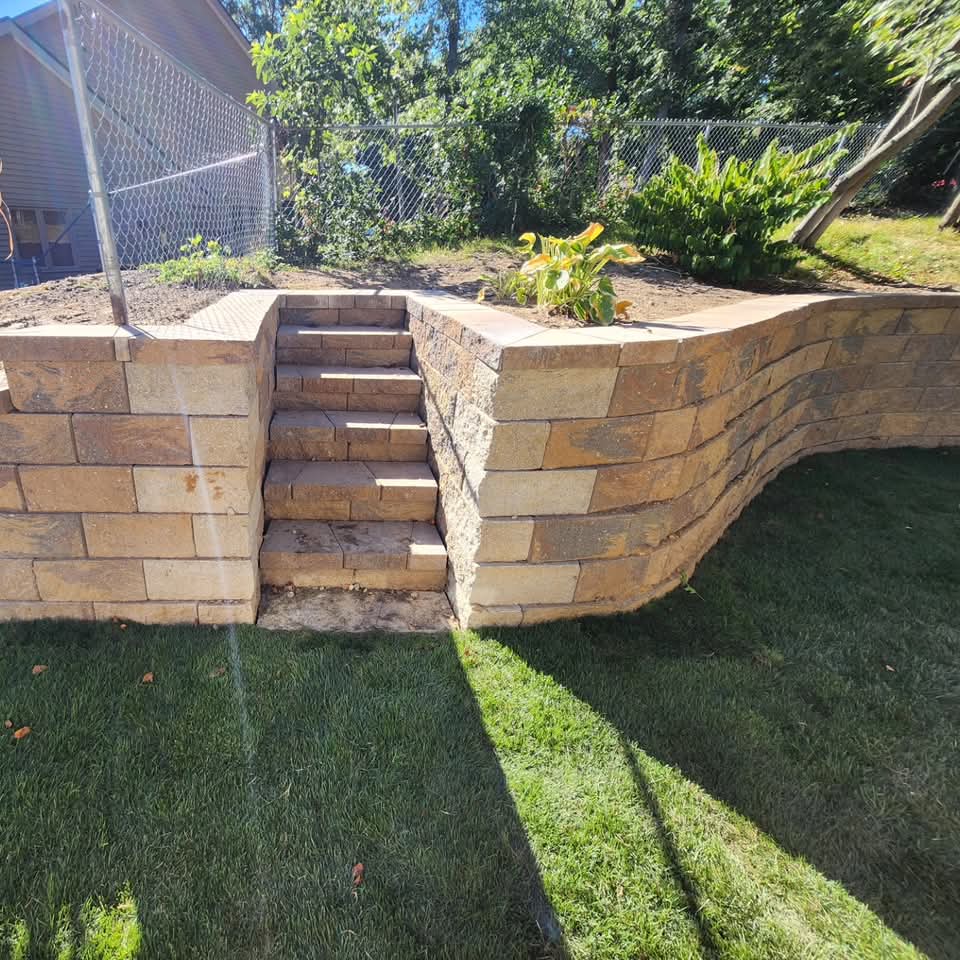Tan block retaining wall with center stairs and a planting bed in a sodded West Michigan backyard