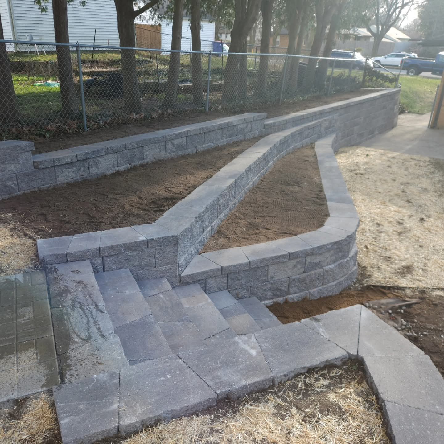 Gray block retaining wall with paver steps and planting beds along a chain-link fence in a front yard
